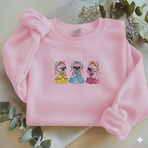 Puede incluir: Una sudadera rosa claro con un dise&ntilde;o bordado de tres personajes de dibujos animados con vestidos de princesa. Los personajes visten vestidos amarillos, azules y rosas, cada uno con una corona. La sudadera tiene mangas largas y cuello redondo.