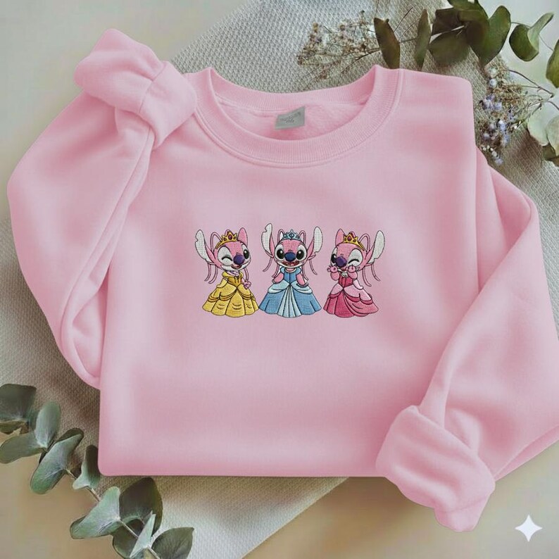 Puede incluir: Una sudadera rosa claro con un dise&ntilde;o bordado de tres personajes de dibujos animados con vestidos de princesa. Los personajes visten vestidos amarillos, azules y rosas, cada uno con una corona. La sudadera tiene mangas largas y cuello redondo.