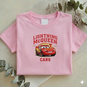 Puede incluir: Una camiseta rosa claro con un dise&ntilde;o bordado de Rayo McQueen de la pel&iacute;cula Cars. El dise&ntilde;o incluye el texto "LIGHTNING McQUEEN" sobre un coche de carreras rojo, con la palabra "CARS" debajo.