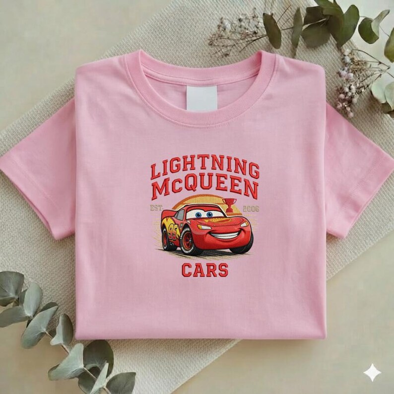 Puede incluir: Una camiseta rosa claro con un dise&ntilde;o bordado de Rayo McQueen de la pel&iacute;cula Cars. El dise&ntilde;o incluye el texto "LIGHTNING McQUEEN" sobre un coche de carreras rojo, con la palabra "CARS" debajo.
