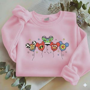 Może przedstawiać: Jasnor&oacute;żowa bluza z kolorową grafiką balon&oacute;w z postaciami z kresk&oacute;wek. Balony, z uszami Myszki Miki, przedstawiają r&oacute;żne postacie. Projekt zawiera gwiazdki i sznurki balon&oacute;w.