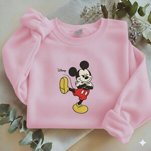 Puede incluir: Sudadera rosa claro con un bordado de Mickey Mouse. El personaje de dibujos animados se muestra en su pose cl&aacute;sica, con pantalones cortos rojos, zapatos amarillos y guantes blancos. El logotipo de Disney est&aacute; impreso en la parte superior izquierda.