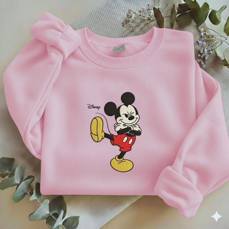 Puede incluir: Sudadera rosa claro con un bordado de Mickey Mouse. El personaje de dibujos animados se muestra en su pose cl&aacute;sica, con pantalones cortos rojos, zapatos amarillos y guantes blancos. El logotipo de Disney est&aacute; impreso en la parte superior izquierda.
