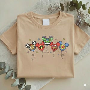 Może przedstawiać: Beżowa koszulka z kr&oacute;tkim rękawem z grafiką balon&oacute;w z postaciami z kresk&oacute;wek. Balony zaprojektowano jako głowy Myszki Miki, z postaciami takimi jak Buzz Lightyear i Zygzak McQueen. Projekt zawiera gwiazdki i sznurki balon&oacute;w.