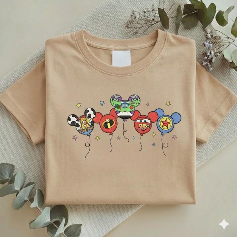 Może przedstawiać: Beżowa koszulka z kr&oacute;tkim rękawem z grafiką balon&oacute;w z postaciami z kresk&oacute;wek. Balony zaprojektowano jako głowy Myszki Miki, z postaciami takimi jak Buzz Lightyear i Zygzak McQueen. Projekt zawiera gwiazdki i sznurki balon&oacute;w.