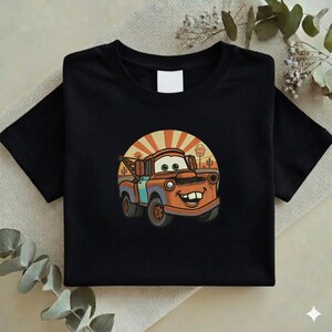 May include: Black t-shirt featuring a cartoon illustration of a smiling, rusty orange tow truck with blue accents. The design includes a desert scene with a setting sun. The shirt is folded.