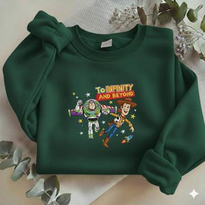 K&ouml;nnte beinhalten: Dunkelgr&uuml;nes Sweatshirt mit einem Aufdruck von Buzz Lightyear und Woody aus Toy Story, mit dem Text "To Infinity and Beyond". Das Design umfasst Sterne und eine kleine Rakete.