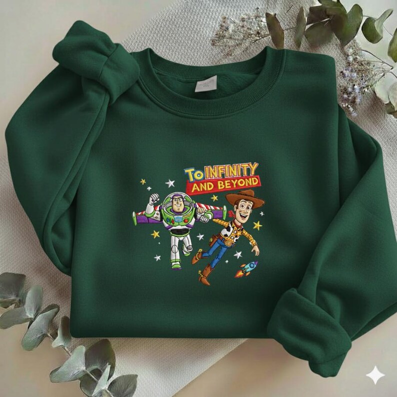 K&ouml;nnte beinhalten: Dunkelgr&uuml;nes Sweatshirt mit einem Aufdruck von Buzz Lightyear und Woody aus Toy Story, mit dem Text "To Infinity and Beyond". Das Design umfasst Sterne und eine kleine Rakete.
