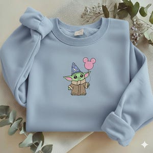 Op de afbeelding: Een lichtblauw sweatshirt met een geborduurd cartoonfiguur dat een roze ballon vasthoudt. Het figuur draagt een tovenaarsmuts en een bruine jas. De sweatshirt heeft een ronde hals en lange mouwen.