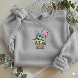 Op de afbeelding: Een lichtgrijs sweatshirt met een geborduurd stripfiguur dat een roze ballon vasthoudt. Het personage is groen met grote oren en draagt een tovenaars hoed. De sweatshirt heeft een ronde hals en lange mouwen.