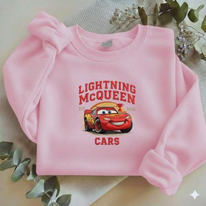 Puede incluir: Una sudadera rosa claro con un dise&ntilde;o de Rayo McQueen de la pel&iacute;cula Cars. El dise&ntilde;o incluye el texto "LIGHTNING MCQUEEN" sobre un coche de carreras rojo, con la palabra "CARS" debajo.