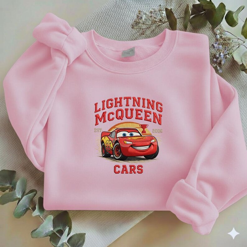 Puede incluir: Una sudadera rosa claro con un dise&ntilde;o de Rayo McQueen de la pel&iacute;cula Cars. El dise&ntilde;o incluye el texto "LIGHTNING MCQUEEN" sobre un coche de carreras rojo, con la palabra "CARS" debajo.