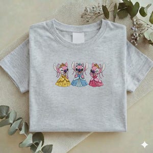 Puede incluir: Una camiseta gris claro con un dise&ntilde;o bordado de tres personajes de dibujos animados vestidos de princesas. Los personajes son rosas con coronas y vestidos en amarillo, azul y rosa. La camiseta est&aacute; doblada cuidadosamente.