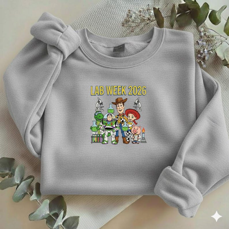 Puede incluir: Una sudadera gris claro con un gr&aacute;fico colorido de personajes de dibujos animados y el texto "LAB WEEK 2026". El gr&aacute;fico incluye un dinosaurio, un guardabosques espacial, un vaquero, una vaquera y un cerdo, frente a equipos de laboratorio.