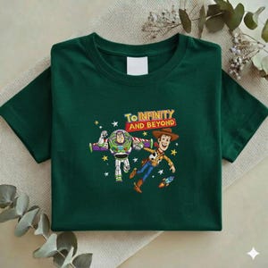 K&ouml;nnte beinhalten: Dunkelgr&uuml;nes T-Shirt mit einer Grafik von Buzz Lightyear und Woody aus Toy Story, mit dem Text "To Infinity and Beyond". Das Design umfasst Sterne und eine Rakete. Das T-Shirt ist gefaltet.