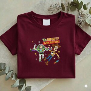 K&ouml;nnte beinhalten: Burgunderfarbenes T-Shirt mit einem farbenfrohen Aufdruck von Buzz Lightyear und Woody aus Toy Story. Der Schriftzug "To Infinity and Beyond" befindet sich &uuml;ber den Figuren. Das Design umfasst Sterne und eine Rakete.