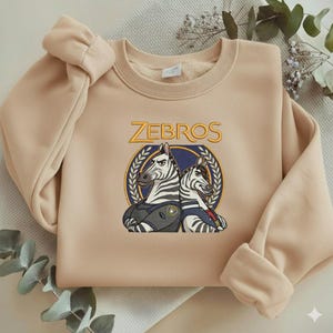 Może przedstawiać: Beżowa bluza z napisem "ZEBROS" w złotych literach nad ilustracją dw&oacute;ch zebr w mundurach. Zebry umieszczone są na niebieskim tle z motywem wieńca laurowego.