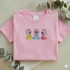 Puede incluir: Una camiseta rosa claro con un dise&ntilde;o bordado de tres personajes de dibujos animados con vestidos de princesa. Los personajes son rosas con coronas y visten vestidos amarillos, azules y rosas. La camiseta est&aacute; doblada sobre una superficie neutra.