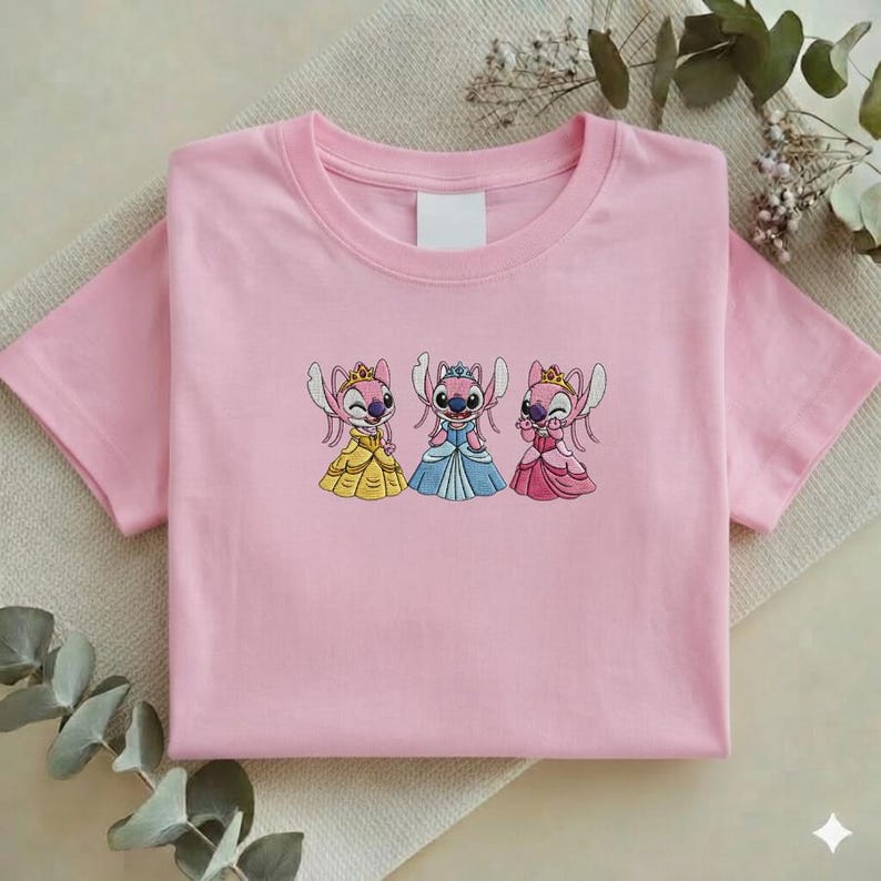 Puede incluir: Una camiseta rosa claro con un dise&ntilde;o bordado de tres personajes de dibujos animados con vestidos de princesa. Los personajes son rosas con coronas y visten vestidos amarillos, azules y rosas. La camiseta est&aacute; doblada sobre una superficie neutra.
