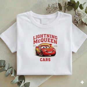 Puede incluir: Camiseta blanca con un dise&ntilde;o de Rayo McQueen de la pel&iacute;cula Cars. El dise&ntilde;o incluye el texto "LIGHTNING MCQUEEN" sobre un coche de carreras rojo sonriente, con "EST. 2006" y la palabra "CARS" debajo.