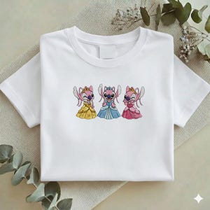 Puede incluir: Camiseta blanca con un dise&ntilde;o bordado de tres personajes de dibujos animados con vestidos de princesa. Los personajes son rosas con coronas y vestidos amarillos, azules y rosas. La camiseta est&aacute; doblada sobre una superficie neutra.
