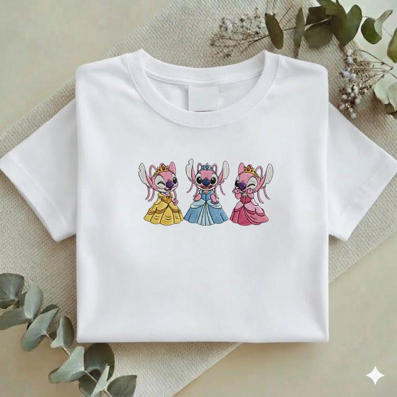 Puede incluir: Camiseta blanca con un dise&ntilde;o bordado de tres personajes de dibujos animados con vestidos de princesa. Los personajes son rosas con coronas y vestidos amarillos, azules y rosas. La camiseta est&aacute; doblada sobre una superficie neutra.
