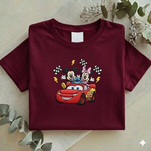 May include: A maroon t-shirt featuring an embroidered design of Mickey Mouse, Minnie Mouse, Stitch, and Lightning McQueen. The design includes checkered flags and lightning bolts. The number 95 is on the car.