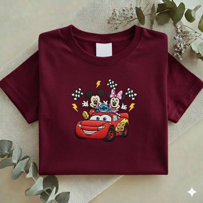 May include: A maroon t-shirt featuring an embroidered design of Mickey Mouse, Minnie Mouse, Stitch, and Lightning McQueen. The design includes checkered flags and lightning bolts. The number 95 is on the car.