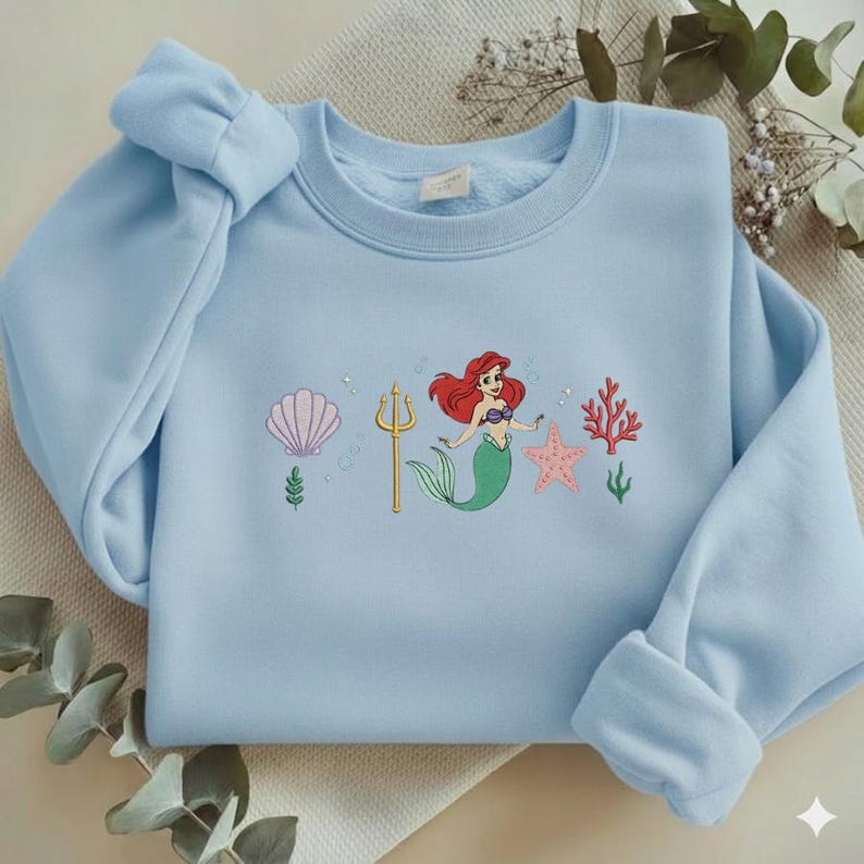 Puede incluir: Sudadera azul claro con un dise&ntilde;o bordado de una sirena con cabello rojo, cola verde y un tridente dorado. El dise&ntilde;o tambi&eacute;n incluye una concha rosa, una estrella de mar rosa y coral rojo. La sudadera tiene cuello redondo y mangas largas.