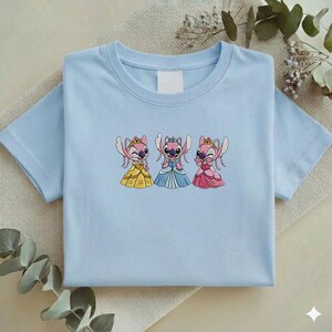 Puede incluir: Camiseta azul claro con un dise&ntilde;o bordado de tres personajes de dibujos animados con vestidos de princesa. Los personajes son rosas con coronas y visten vestidos amarillos, azules y rosas. La camiseta est&aacute; doblada cuidadosamente.