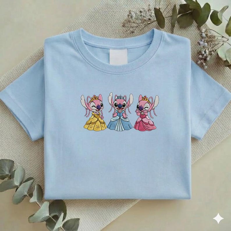 Puede incluir: Camiseta azul claro con un dise&ntilde;o bordado de tres personajes de dibujos animados con vestidos de princesa. Los personajes son rosas con coronas y visten vestidos amarillos, azules y rosas. La camiseta est&aacute; doblada cuidadosamente.