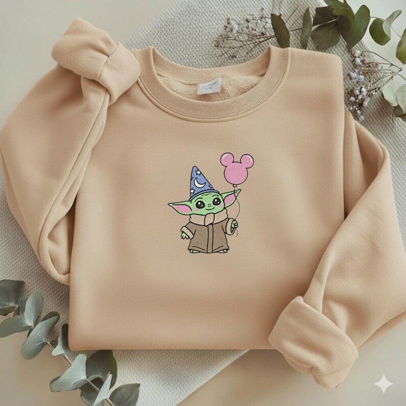 Op de afbeelding: Een beige sweatshirt met een geborduurd stripfiguur met een tovenaars hoed en een roze ballon. Het personage is groen met grote ogen en draagt een bruine jas. De sweatshirt heeft een ronde hals en lange mouwen met geribde manchetten.