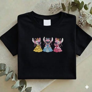 Puede incluir: Camiseta negra con un dise&ntilde;o bordado de tres personajes de dibujos animados vestidos de princesas. Cada personaje lleva una corona y un vestido de diferente color: amarillo, azul y rosa. La camiseta est&aacute; doblada cuidadosamente.