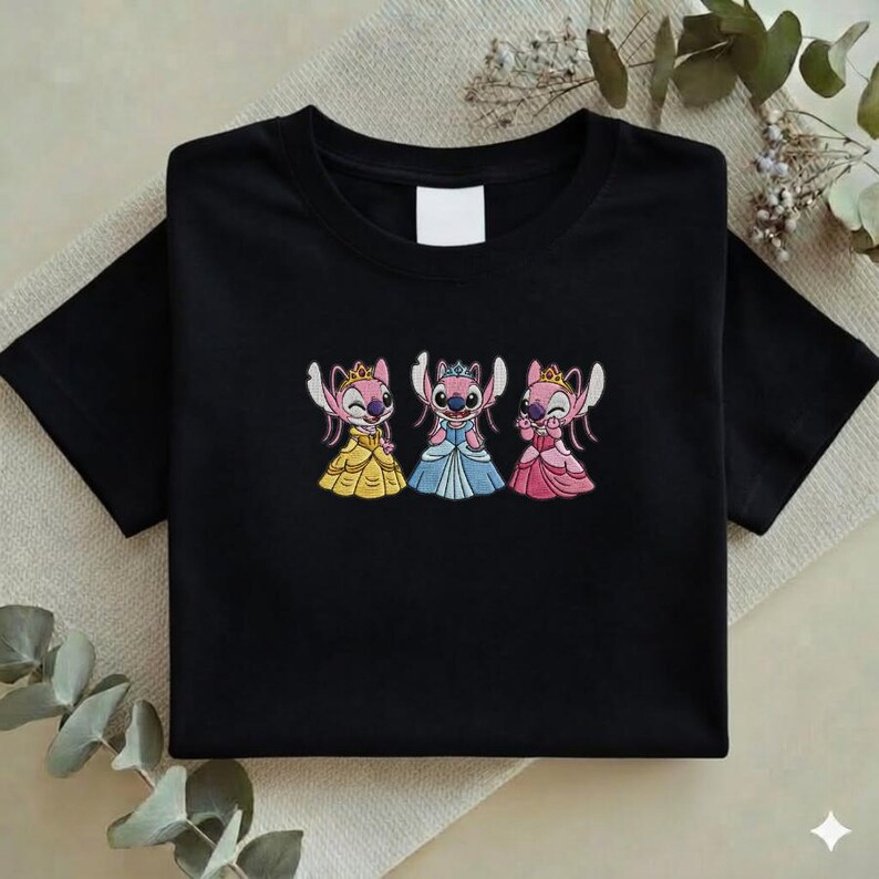 Puede incluir: Camiseta negra con un dise&ntilde;o bordado de tres personajes de dibujos animados vestidos de princesas. Cada personaje lleva una corona y un vestido de diferente color: amarillo, azul y rosa. La camiseta est&aacute; doblada cuidadosamente.