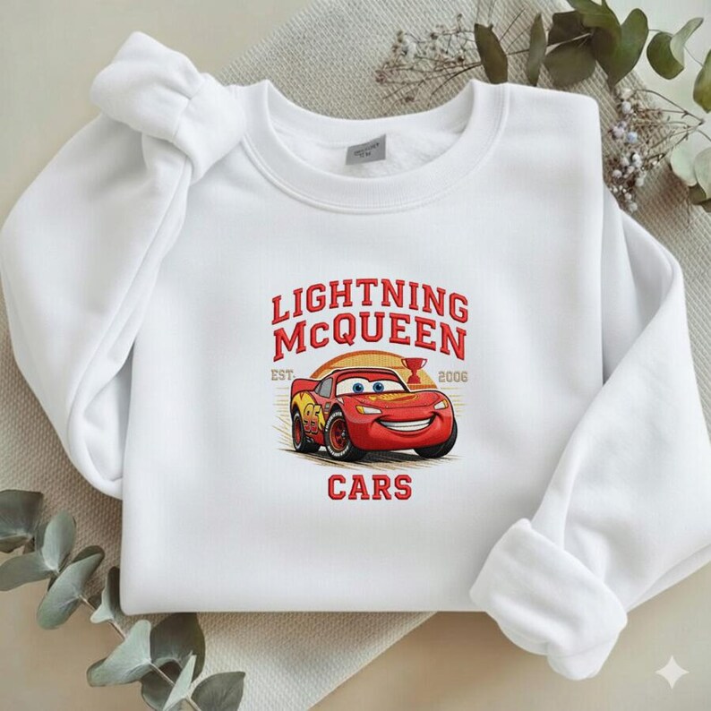 Puede incluir: Sudadera blanca con un dise&ntilde;o de Lightning McQueen de la pel&iacute;cula Cars. El dise&ntilde;o incluye el texto "LIGHTNING MCQUEEN" en rojo, con "EST. 2006" y "CARS" debajo de la imagen del coche. La sudadera tiene cuello redondo y mangas largas.