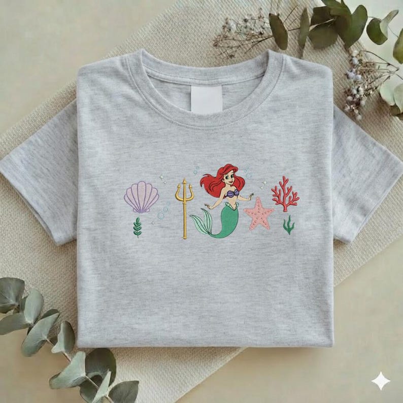 Puede incluir: Camiseta gris claro con un dise&ntilde;o bordado. El dise&ntilde;o presenta una sirena con cabello rojo y cola verde, una concha, un tridente, una estrella de mar y coral. El dise&ntilde;o es en rosa, verde y dorado.