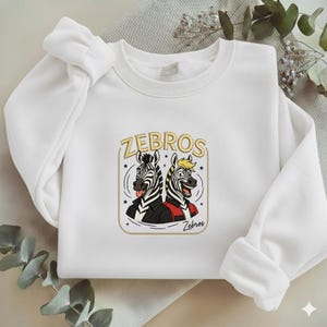 May include: White sweatshirt featuring a graphic of two zebras with the text "ZEBROS". One zebra is wearing a black jacket, the other a red jacket. The design is in a gold frame.