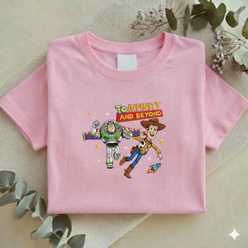 K&ouml;nnte beinhalten: Ein hellrosa T-Shirt mit einer Grafik von Buzz Lightyear und Woody aus Toy Story, mit dem Text "To Infinity and Beyond". Das Design umfasst Sterne und eine kleine Rakete. Das T-Shirt ist auf einer neutralfarbenen Oberfl&auml;che gefaltet.
