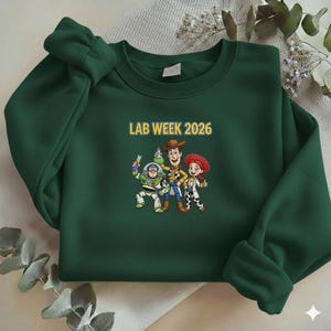 May include: A dark green sweatshirt with the text "LAB WEEK 2026" in yellow. The sweatshirt features a cartoon graphic of three characters. The characters are Buzz Lightyear, Woody, and Jessie.