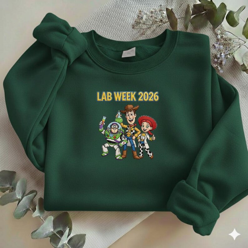 May include: A dark green sweatshirt with the text "LAB WEEK 2026" in yellow. The sweatshirt features a cartoon graphic of three characters. The characters are Buzz Lightyear, Woody, and Jessie.