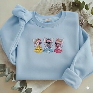 Puede incluir: Sudadera azul claro con un bordado de tres personajes de dibujos animados con vestidos de princesa. Los personajes son rosas y llevan coronas, con vestidos amarillos, azules y rosas. La sudadera tiene cuello redondo y mangas largas.