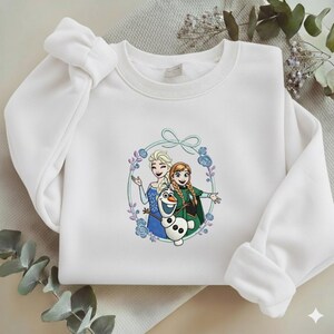 Puede incluir: Sudadera blanca con un gr&aacute;fico colorido de Elsa, Anna y Olaf de Frozen, rodeados por una corona floral. La parte superior de manga larga tiene cuello redondo y un corte holgado.