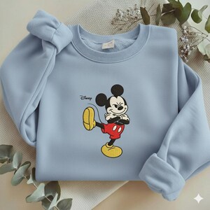 Puede incluir: Sudadera azul claro con un dise&ntilde;o bordado de Mickey Mouse. El personaje de Disney est&aacute; representado con pantalones cortos rojos, zapatos amarillos y orejas negras, con el logotipo de Disney arriba. La sudadera tiene cuello redondo y pu&ntilde;os.
