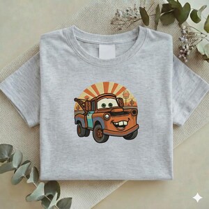 May include: A light gray t-shirt featuring a cartoon image of Mater from the movie Cars. The image shows Mater with a smiling face, set against a desert backdrop with a rising sun. The shirt is folded.