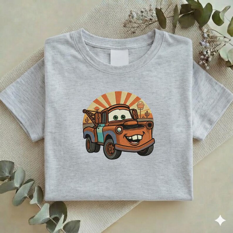 May include: A light gray t-shirt featuring a cartoon image of Mater from the movie Cars. The image shows Mater with a smiling face, set against a desert backdrop with a rising sun. The shirt is folded.