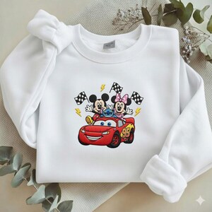 May include: White sweatshirt featuring an embroidered design of Mickey Mouse, Minnie Mouse, and Stitch riding in a red race car. The design includes checkered flags and lightning bolts. The car has the number 95 on the side.