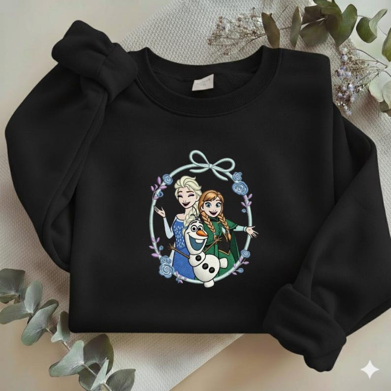 Puede incluir: Sudadera negra con un gr&aacute;fico colorido de Elsa, Anna y Olaf de la pel&iacute;cula Frozen, rodeados por una corona floral. El dise&ntilde;o est&aacute; centrado en el pecho de la parte superior de manga larga.