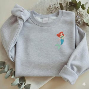 May include: A light gray sweatshirt featuring a small embroidered image of a mermaid with red hair, a green top, and a blue tail. The sweatshirt has long sleeves and a crew neck. The sleeves are folded up at the cuffs.