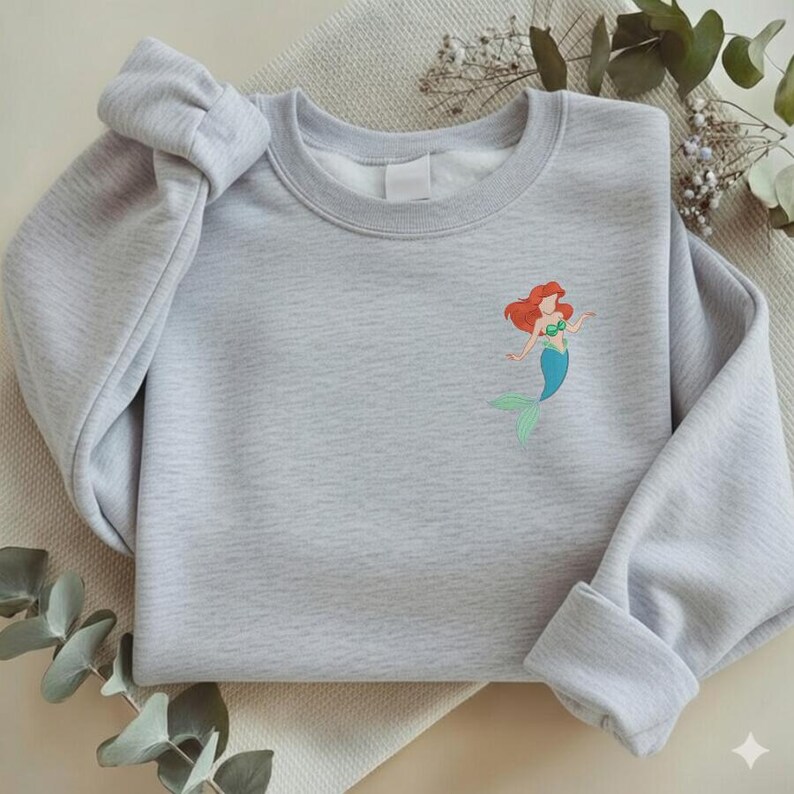 May include: A light gray sweatshirt featuring a small embroidered image of a mermaid with red hair, a green top, and a blue tail. The sweatshirt has long sleeves and a crew neck. The sleeves are folded up at the cuffs.