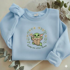 May include: Light blue sweatshirt featuring a cartoon character holding flowers, surrounded by a floral wreath. The character is green with large ears and a tan coat. The text "Enjoy the little things" is below the character.